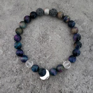 May include: A dark blue and purple gemstone bracelet with a silver crescent moon charm. The bracelet is made with lava rock and clear quartz beads.