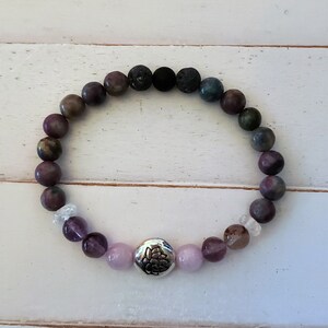 May include: A purple, green, and black beaded bracelet with a silver charm featuring a floral design. The bracelet is made with natural stones and is designed to be worn on the wrist.