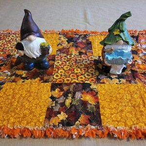 May include: Two garden gnomes on a fall-themed table runner. The gnomes have brown and green hats, white beards, and hold items. The runner is patchwork with orange, yellow, and brown leaf and floral patterns, and orange fringe.