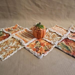 May include: A fall-themed table runner with a patchwork design featuring autumn leaves and a small pumpkin-shaped container. The runner is made of fabric with orange, brown, and cream colors. The pumpkin container is orange with a green stem.