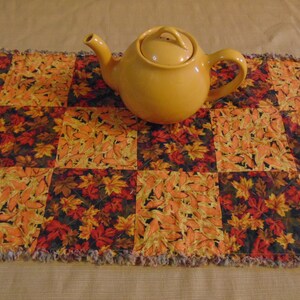 May include: A yellow ceramic teapot sits on a fall-themed table runner. The runner features a patchwork design with autumn leaf patterns in shades of orange, yellow, and red. The edges of the runner have a fringed trim, adding texture to the design.