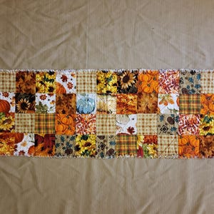 Op de afbeelding: Een rechthoekige patchwork tafelloper met een verscheidenheid aan herfstthema stoffen. Het ontwerp bevat vierkanten met zonnebloemen, pompoenen, herfstbladeren en ruitpatronen. Het kleurenpalet wordt gedomineerd door warme tinten oranje, geel en bruin.