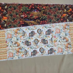 May include: A rectangular rag quilt table runner featuring two distinct fabric patterns. The top section displays turkeys, pumpkins, and a farm scene on a dark brown background. The bottom section shows turkeys and pumpkins on a cream background.