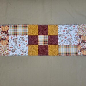 May include: A rectangular quilted table runner featuring a patchwork design. The fabric panels display patterns of orange pumpkins, brown and white hedgehogs, autumn leaves, and a plaid print in shades of yellow, orange, brown, and maroon.