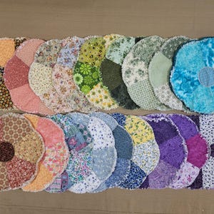May include: A collection of handmade, flower-shaped fabric coasters. Each coaster is made from various patterned fabrics in a range of colors, including floral prints, solid colors, and textured designs. The coasters have a fringed edge.