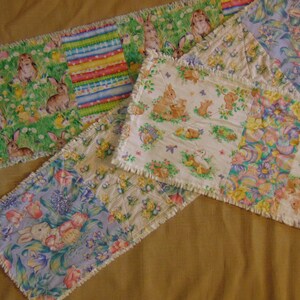 Op de afbeelding: Een verzameling stoffen tafellopers met Pasen als thema. De lopers hebben patchwork-ontwerpen met verschillende patronen, waaronder konijnen, kuikens en bloemmotieven. De randen hebben een gerafelde, onafgewerkte uitstraling en het kleurenpalet omvat pasteltinten en groen.