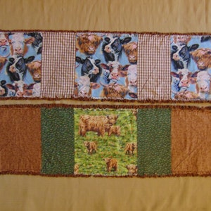 May include: A brown and green patchwork table runner with a brown, green, and white cow print design. The runner is made of fabric and has a fringed edge.