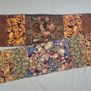 May include: Two rectangular fabric table runners featuring autumn-themed patterns. The top runner displays pumpkins, sunflowers, and leaves in orange, brown, and black. The bottom runner features a patchwork design of leaves, grapes, and gourds.