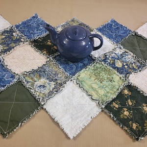 Peut inclure: Une théière bleue est posée sur un chemin de table matelassé. Le chemin de table est composé de différents tissus à motifs bleus, verts et blancs, disposés en losanges. Les bords du chemin de table ont une finition effilochée et texturée.