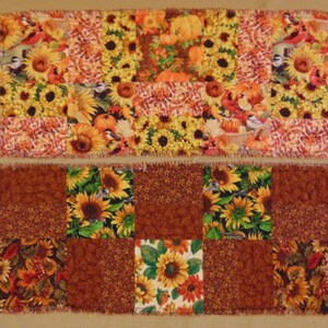 May include: Two rectangular patchwork fabric pieces. The top piece has a pattern of sunflowers, pumpkins, and birds in yellow, orange, and brown. The bottom piece has a similar patchwork design with sunflowers and brown fabric squares.