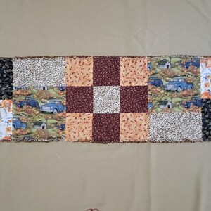 May include: A rectangular rag quilt table runner featuring a patchwork design with various fabric patterns including black leaf prints, orange pumpkins, blue trucks, and brown and tan checkered squares with frayed, exposed seams throughout.