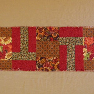 May include: A rectangular patchwork table runner featuring a fall theme. The runner is composed of various fabric squares in shades of red, brown, green, and yellow. The design includes floral and leaf patterns, with a frayed edge.