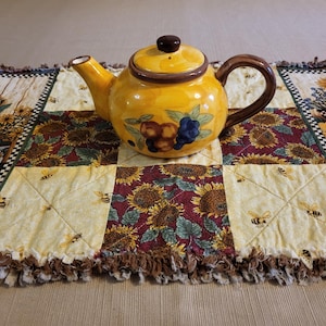 May include: A yellow ceramic teapot with a brown handle and lid, decorated with fruit and floral designs. The teapot sits on a quilted table runner with sunflowers, bees, and cornucopia patterns.