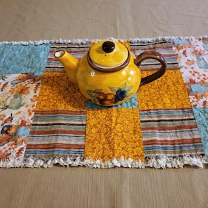 May include: A yellow ceramic teapot with a brown handle and lid sits on a patchwork table runner. The runner features squares of blue, orange, and white fabric with floral and leaf patterns. The teapot has a floral design.