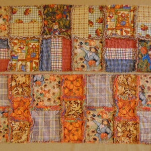 May include: A patchwork textile featuring squares of fabric with autumn-themed patterns. The design includes images of scarecrows, pumpkins, sunflowers, and plaid patterns in shades of orange, brown, and blue. The edges of the squares have a frayed, rag-quilt style.