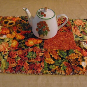 May include: A white ceramic teapot with a floral design sits on a fall-themed table runner. The runner features a patchwork design with pumpkins, sunflowers, and autumn leaves in shades of orange, red, and yellow.