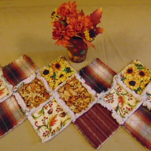 May include: A handmade, patchwork table runner featuring a repeating diamond pattern with autumn-themed fabrics. The fabrics include sunflowers, fall leaves, and plaid patterns in shades of red, orange, and yellow. A small vase of fall flowers sits in the background.