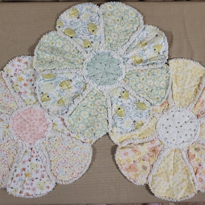May include: Three handmade, flower-shaped rag quilts. Each quilt features a different floral pattern in pastel colors, including pink, yellow, and green. The quilts have a ruffled edge and a textured appearance.