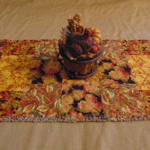 May include: A fall-themed table runner with a patchwork design featuring autumn leaves in shades of orange, yellow, and brown. A decorative turkey figurine sits atop the runner, adding to the seasonal decor. The runner is approximately 12 inches wide.