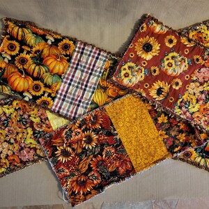 May include: A collection of rectangular fabric placemats featuring autumn-themed patterns including orange pumpkins, yellow sunflowers, and fall foliage. The pieces are constructed with frayed, exposed seams and incorporate various plaid and floral prints.