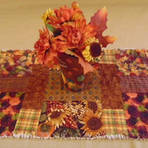 May include: A fall-themed table runner with a patchwork design featuring sunflowers, leaves, and plaid patterns in shades of orange, brown, and green. A small decorative vase with orange flowers and leaves sits on top.