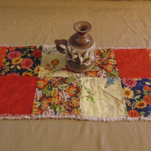 May include: A patchwork table runner with a variety of floral and solid red squares. A brown ceramic vase with a handle and floral accents sits on the runner. The runner has a fringed edge.