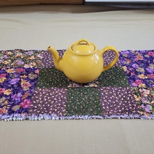 Può includere: Una teiera in ceramica giallo brillante con corpo arrotondato, beccuccio curvo e coperchio, centrata su un runner da tavola trapuntato rettangolare caratterizzato da un motivo patchwork di quadrati in tessuto floreale viola, verde e oro.