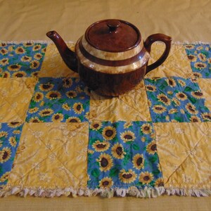 May include: A brown teapot with a white stripe sits on a yellow and blue quilted table runner with a sunflower pattern.