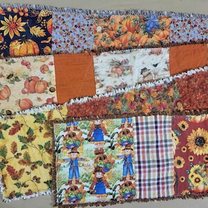 Può includere: Un runner da tavola rettangolare in stile rag quilt con un design patchwork realizzato con vari tessuti a tema autunnale. I motivi includono zucche, girasoli, foglie autunnali, spaventapasseri e quadri nei toni dell'arancione, giallo, marrone e blu navy.