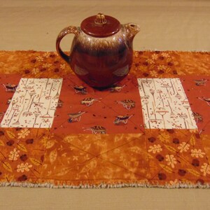 May include: A brown ceramic teapot with a textured surface sits on a quilted table runner. The runner features a patchwork design with orange, brown, and cream fabrics. The pattern includes wheelbarrows, rakes, and leaf motifs.