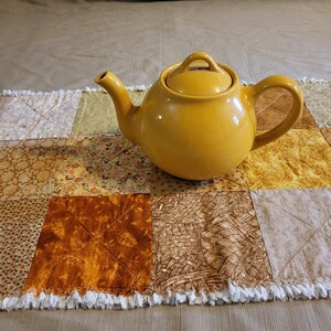 May include: A mustard-yellow ceramic teapot with a lid rests on a patchwork quilt runner. The runner is made of squares in gold, brown, and cream, with a fringed edge. The teapot has a curved handle and spout, perfect for serving tea.