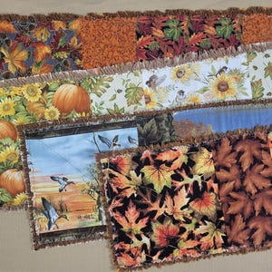 May include: A collection of rectangular fabric table runners featuring various autumn-themed patterns including orange pumpkins, yellow sunflowers, flying ducks over a landscape, and multicolored maple leaves, all finished with a frayed edge.