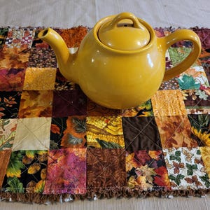 May include: A yellow ceramic teapot with a lid sits on a patchwork table runner. The runner features a variety of autumn-themed fabrics in shades of orange, brown, yellow, and green. The teapot has a curved handle and spout.