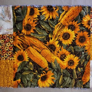 May include: A patchwork table runner with a central panel of sunflowers and corn on a black background. The design is framed by panels with floral and wheat patterns in yellow, orange, and cream. The edges have a frayed finish.