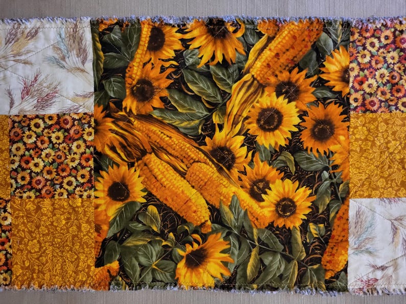 May include: A patchwork table runner with a central panel of sunflowers and corn on a black background. The design is framed by panels with floral and wheat patterns in yellow, orange, and cream. The edges have a frayed finish.