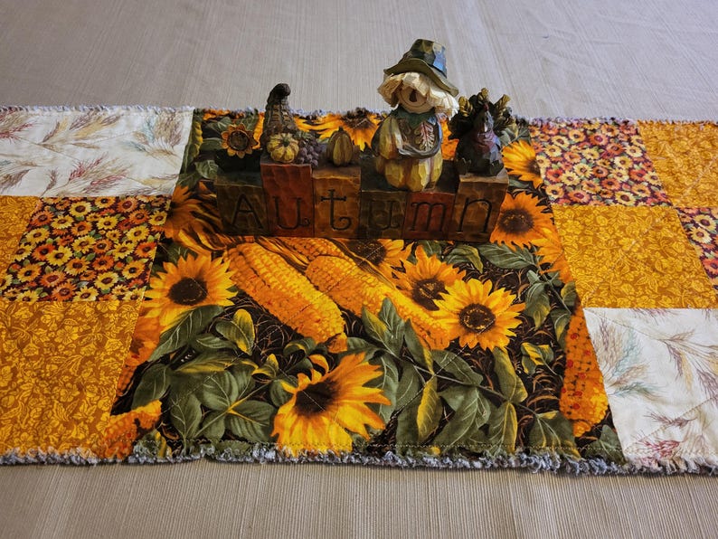 May include: A patchwork table runner with a design of sunflowers, corn, and autumn figurines. The word "AUTUMN" is displayed on a wooden block. The runner has a frayed edge. The measurements are in centimetres.