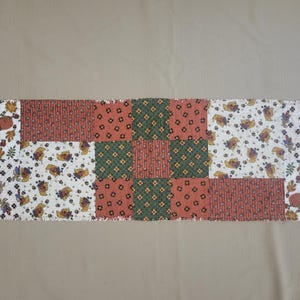 May include: A rectangular fabric table runner featuring a patchwork design of alternating squares. The pattern includes white fabric with autumn leaves and pumpkins, red fabric with thin vertical stripes, and green fabric with small floral motifs.