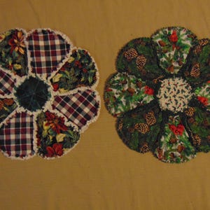 May include: Two flower-shaped quilted trivets. One features plaid and floral patterns in red, green, and white. The other has a green pinecone and red berry design. Both have a white, fringed edge.