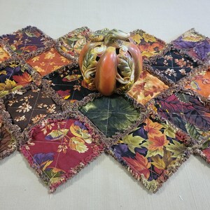 May include: A fall-themed table runner with a patchwork design featuring leaf patterns in shades of orange, burgundy, green, and purple. A ceramic pumpkin decorates the center of the runner, adding to the autumn aesthetic.