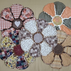 May include: A collection of handmade, rag-style flower-shaped trivets. Each trivet features a unique patchwork design with various patterns and colors, including gingham, floral, and polka dots. The trivets are designed to protect surfaces from heat.