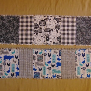 May include: A farmhouse-themed table runner with a patchwork design. The runner is made of different fabrics with black and white checked patterns, a black and white farm animal print, and a blue and white farm animal print. The runner has a frayed edge.