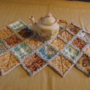 May include: A patchwork quilted tea cozy with a blue and brown color scheme. The cozy is made of squares of fabric with a frayed edge. A white teapot with floral designs sits on top of the cozy.
