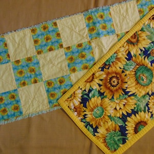 May include: Two decorative table runners. One features a patchwork design with yellow squares and sunflower fabric. The other has a blue background with large sunflowers and a yellow border. Both are quilted.
