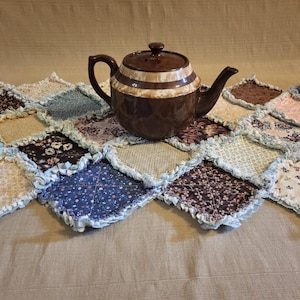 May include: A brown ceramic teapot with cream-colored stripes sits on a patchwork quilt. The quilt is made of various patterned fabrics in shades of blue, brown, and cream. The teapot has a lid and a handle.