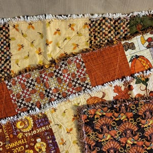 Op de afbeelding: Een patchwork textiel met herfstthema's. Het ontwerp omvat vierkanten met pompoenen, bladeren, kalkoenen en ruitpatronen in oranje, bruin en geel. De randen hebben een gefranjerde rand.