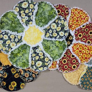 Può includere: Una collezione di oggetti in tessuto fatti a mano a forma di fiore. I petali presentano vari motivi di girasoli in nero, verde, rosso e giallo. Il centro del fiore più grande è giallo. I bordi sono rifiniti con frange bianche.