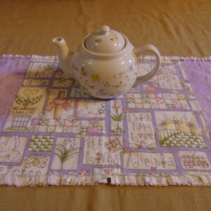 Può includere: Una teiera bianca con motivi floreali si trova su un runner da tavolo trapuntato viola e bianco con un bordo a frangia. Il runner ha un design a patchwork con varie frasi tra cui "Faith Hope Love" e "Alleluia".
