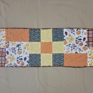May include: A rectangular rag quilt table runner featuring a patchwork design of squares in various patterns including orange polka dots, yellow floral prints, dark blue textured fabric, white fabric with autumn leaves, and brown plaid fabric.