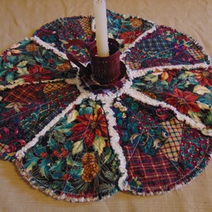 Könnte beinhalten: Ein handgefertigtes, gestepptes Kerzen-Pad mit Blumen- und Karomuster. Die Matte hat die Form einer Blume mit einem weißen, gerüschten Rand. Ein burgunderfarbener Kerzenhalter aus Metall hält eine weiße Kerze.