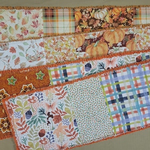 May include: A rectangular rag quilt table runner featuring a patchwork design of various autumn-themed fabrics. The patterns include orange pumpkins, colorful fall leaves, acorns, mushrooms, butterflies, and plaid prints in shades of orange, brown, and blue.